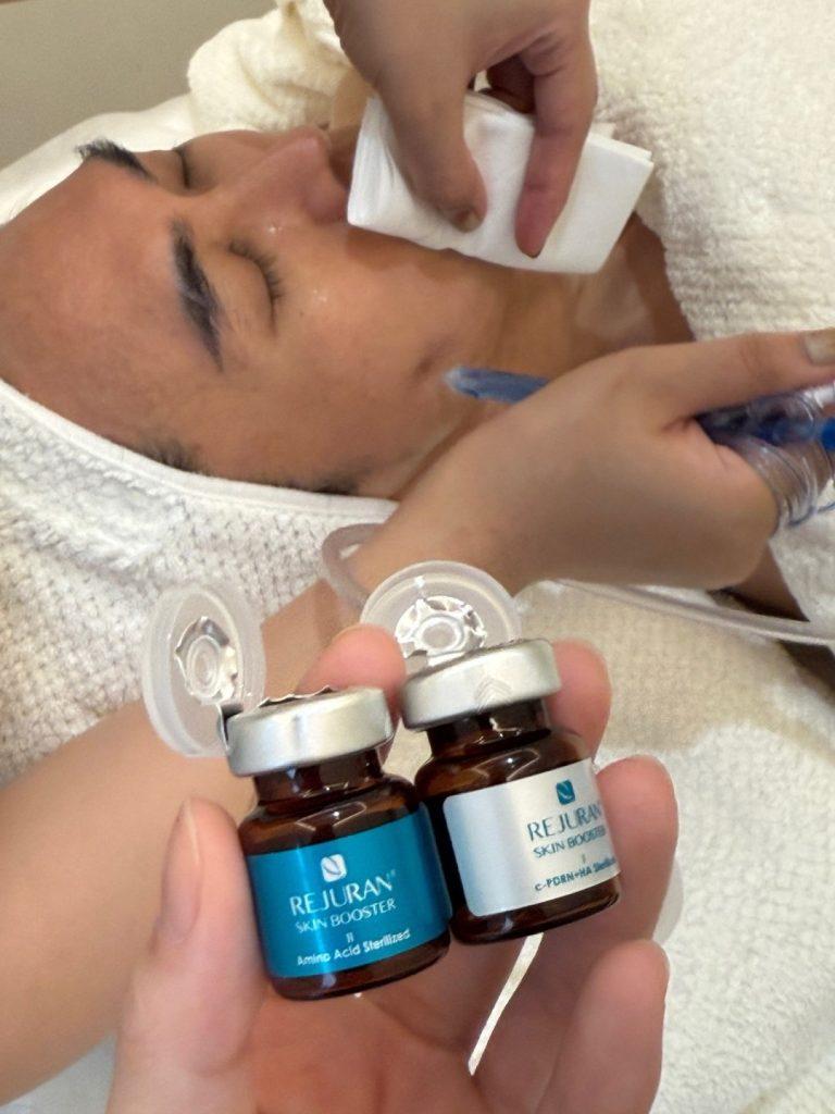 兩瓶 Rejuran Skin Booster 的特寫:一瓶胺基酸滅菌,另一瓶 C-PDRN-HA 滅菌。背景中,一位女士正在接受臉部護理。